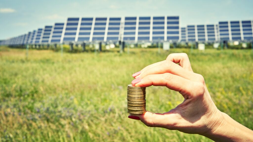 Mano sosteniendo columna de monedas con campo fotovoltaico al fondo; Subvenciones placas solares vitoria-gasteiz, Álava, País Vasco