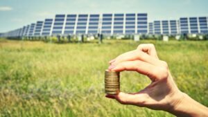 Mano sosteniendo columna de monedas con campo fotovoltaico al fondo; Subvenciones placas solares vitoria-gasteiz, Álava, País Vasco